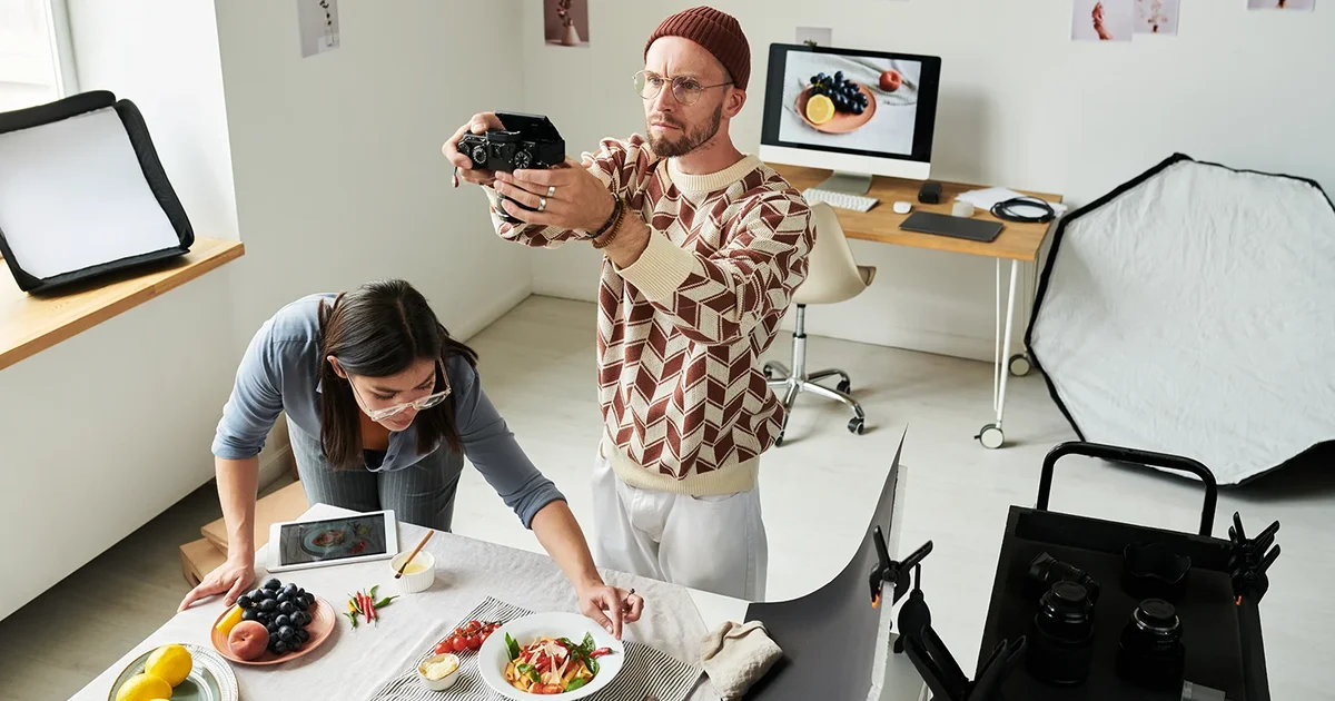 Professional food photographer and stylist preparing a dish in studio with lighting equipment, camera on tripod, and computer monitor showing food images - large